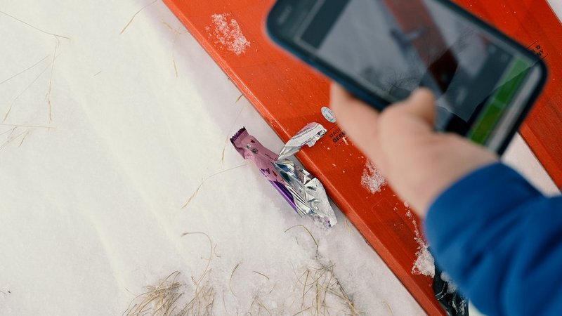 Person auf Skiern macht Foto mit Smartphone von Plastikmüll auf Schnee