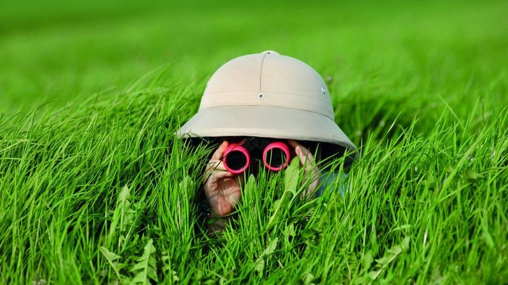 Person liegt im hohen Gras mit Safari-Hut und beobachtet durch Ferngläser